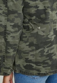 Camouflage jacket in varying shades of green and black, featuring metallic snap detail and a side pocket, made from textured fabric.