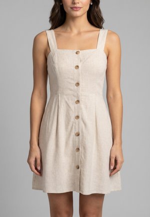 Mujer con vestido beige sin mangas con botones delanteros, tirantes anchos y escote cuadrado sobre un fondo gris.