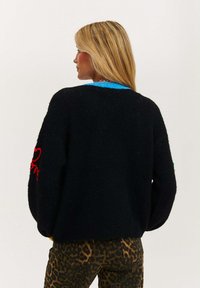 Czarny sweter dzianinowy o luźnym kroju, z niebieskim kołnierzykiem, czerwonym haftem na rękawach i teksturowanym wykończeniem. Połączony z spodniami w panterkę.