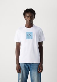 Jaunas vyras su trumpais susisukusiais plaukais, vilkintis baltą Calvin Klein marškinėlį su mėlynu logotipu ir šviesiai mėlynomis suplėšytomis džinsomis prieš baltą foną.