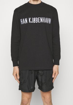 Personne portant un t-shirt noir à manches longues avec un texte blanc "HAN KJØBENHAVN" et un short noir sur un fond clair uni.