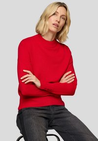 Roter gestrickter Pullover mit hohem Kragen und gerippten Bündchen, verziert mit Knopfdetails an den Ärmeln. Kombiniert mit dunkelgrauen Jeans.