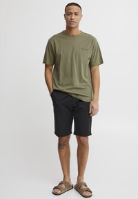 Olive green cotton t-shirt with a chest pocket, paired with black shorts. The model wears brown sandals and has a tattoo visible on his leg.