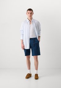 Chemise à rayures bleu clair sur un débardeur blanc, associée à un bermuda cargo bleu marine et des chaussures slip-on marron. Chaussettes blanches visibles.
