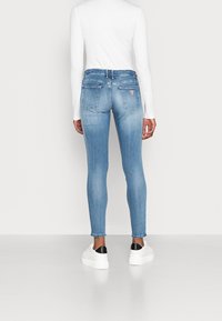 Jeans ajustados de mezclilla azul claro, de cintura alta con bolsillos traseros, combinados con una blusa blanca de manga larga y zapatillas blancas con detalles en negro.