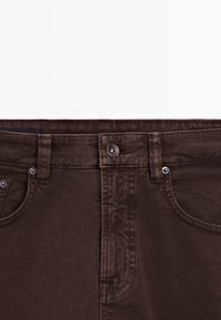Pantalon en jean brun foncé en gros plan montrant le bouton avant, les passants de ceinture et les poches avant sur un fond blanc uni.