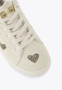 Beżowe zamszowe sneakersy z płaskimi sznurowadłami, złotym akcentem na języku i dwoma łatkami w kształcie serca z fakturowanym wzorem po bokach.