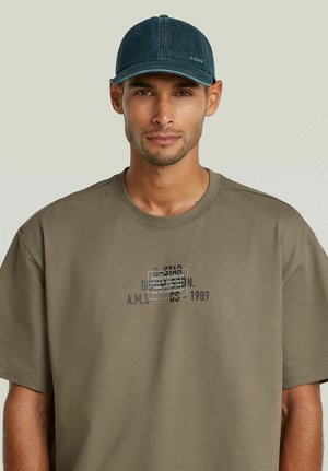 Uomo che indossa una t-shirt verde oliva con stampa e un cappellino blu scuro, rivolto in avanti su uno sfondo chiaro uniforme.