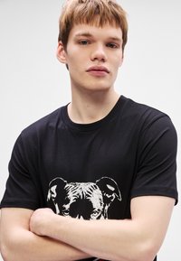 T-shirt preta com um gráfico branco da face de um cão. Mangas curtas, decote redondo, feita de um material suave com uma textura lisa.