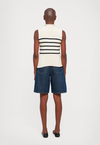 Maglione senza maniche di colore crema con strisce orizzontali nere, abbinato a pantaloni corti in denim scuro e scarpe slip-on nere, visto da dietro.