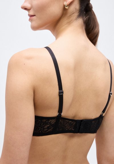 Γυναίκα που φοράει μαύρο δαντελένιο σουτιέν με ρυθμιζόμενους ιμάντες και χρυσό σκουλαρίκι σε σχήμα κρίκου, εμφανίζεται από πίσω και πλάγια προφίλ μπροστά σε λευκό φόντο.