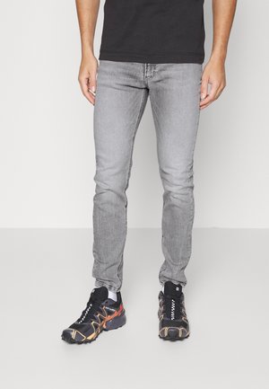 Man draagt slanke grijze spijkerbroek en zwarte sportschoenen met oranje en witte accenten, staande tegen een effen achtergrond.