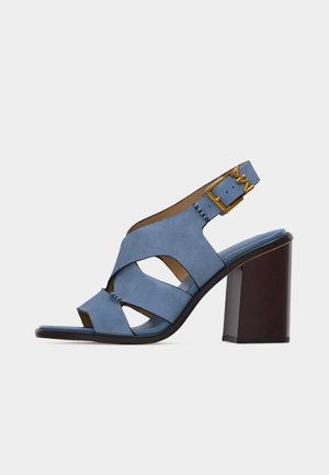 Sandale ouverte à talon en daim bleu avec larges brides croisées, boucle dorée et talon bloc épais marron foncé sur fond uni.