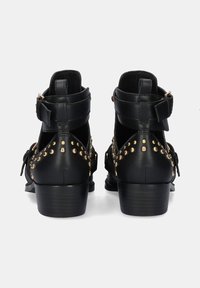 Botas de cuero negras hasta el tobillo con un tacón bajo, que presentan tachuelas doradas y una correa ajustable en el tobillo. Textura suave con detalles de encaje.