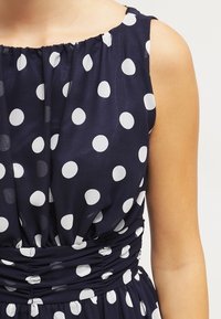 Marineblaues Kleid mit weißen Punkten, das eine geraffte Halslinie und einen elastischen Bund für eine figurbetonte Passform aufweist. Glattes Gewebe mit weicher Textur.