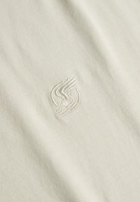 Tissu en coton crème avec un logo brodé en relief subtil présentant un motif d'ailes délicat en fil léger. Texture douce, finition lisse.