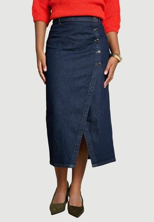 Mujer con una falda midi de mezclilla azul oscuro con botones a lo largo de la abertura frontal, combinada con un suéter rojo y zapatos oscuros puntiagudos.