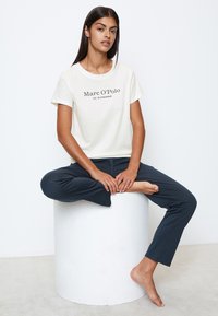 Creme t-shirt med sort tryk "Marc O'Polo EST. I STOCKHOLM," parret med mørk navy bukser; model siddende på en hvid cylindrisk pedestal.