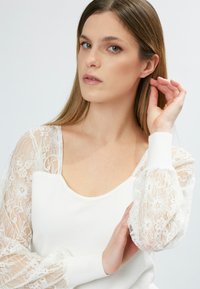 Weißer Pullover mit langen, durchsichtigen Spitzenärmeln, die mit floralen Mustern verziert sind. Die Passform ist tailliert mit einem runden Ausschnitt und gerippten Bündchen.