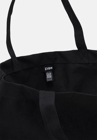 Svart tygkasse med två axelband, med en inre etikett. Designen är enkel utan synliga mönster eller dekorationer.