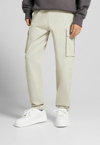 Beiga cargobyxor i bomull med sidofickor och rak benform, kombinerade med vita sneakers och en grå tröja.