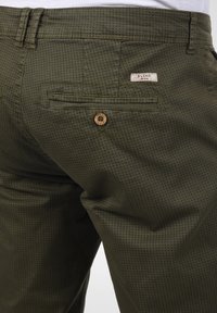 Grønne mønstrede bukser med et struktureret stof, der har en baglomme og en knap i træ. Subtil krysshatch-design.