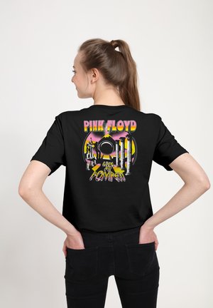 Ung kvinde med hestehale iført sort Pink Floyd "Live at Pompeii" t-shirt med søjler og solnedgangsgrafik på bagsiden.