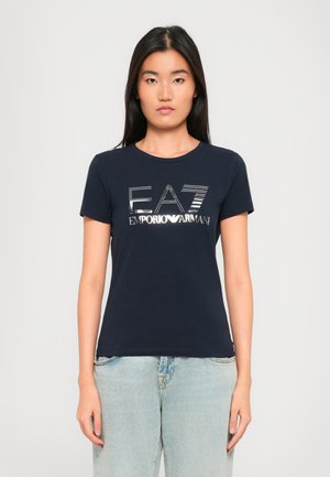 Vrouw met lang donker haar, gekleed in een marineblauwe EA7 Emporio Armani T-shirt en lichtblauwe jeans, staande tegen een effen achtergrond.