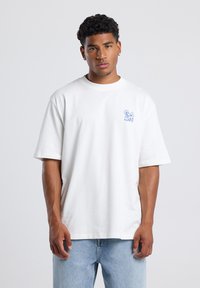White cotton T-shirt with a crew neck and short sleeves, featuring a blue graphic on the left chest. Worn with light blue denim jeans.