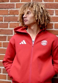 Felpa rossa con zip realizzata in tessuto morbido, dotata di cappuccio, tasche frontali, logo Adidas bianco ed emblema del Bayern Monaco sul petto.
