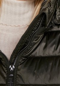 Donkergroene gewatteerde jas met een ritssluiting, met een gladde textuur. Daaronder is een lichtbeige geribbelde trui gedeeltelijk zichtbaar.