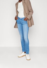 Vaqueros pitillo azul claro de talle alto, combinados con una camiseta blanca y una blazer beige. Zapatillas blancas con suela gruesa completan el conjunto.