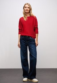 Roter Pullover mit langen Ärmeln, kombiniert mit dunkelblauen weit geschnittenen Jeans und weißen Sneakers, mit einem lockeren Sitz und minimalen Details.