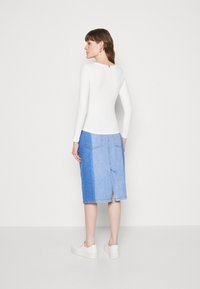 Weißes Langarmoberteil kombiniert mit einem hellblauen Denimrock, der einen Schlitz hinten, seitliche Taschen und eine legere Passform aufweist. Weiße Slipper.