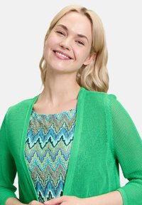 Grüne Netzstrickjacke mit offenem Vorderteil und Dreiviertelärmeln. Darunter ein gemusterter blau-grüner Top mit wellenförmigen Linien.