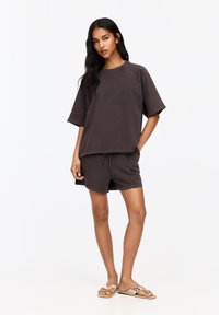 Donkerbruin oversized katoenen t-shirt met korte mouwen, gecombineerd met bijpassende short met trekkoord. Draagt beige sandalen met versieringen.