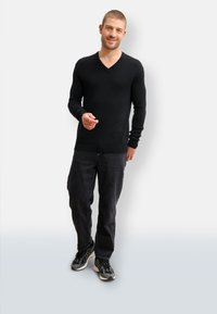 Suéter negro ajustado con cuello en V, de manga larga y tela texturizada, combinado con jeans oscuros y zapatillas negras. El modelo posó frente a un fondo blanco.