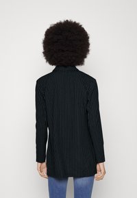 Person med krulligt hår som bär en svart randig kavaj och blå jeans, stående med ansiktet vänt mot en bakgrund i rent vitt.