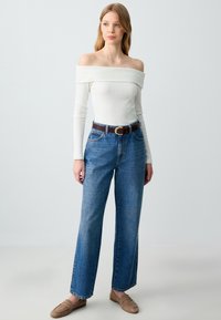 Off-Shoulder-Top aus weißem Rippstoff, kombiniert mit high-waisted blauen Jeans und einem braunen Gürtel. Weiche Textur, lockere Passform und weiter Schnitt.
