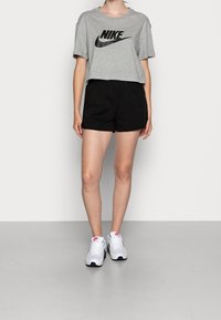 Šedé zkrácené tričko s černým logem "Nike", v kombinaci s černými šortkami. Model má na sobě bílé sportovní boty s modrými a červenými akcenty.