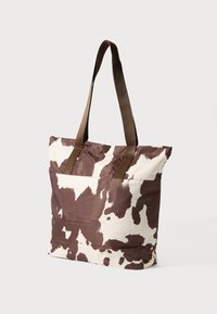 Sac fourre-tout avec un motif imprimé vache en marron et crème, fabriqué en tissu avec des anses marron résistantes. Présente une forme simple et rectangulaire.