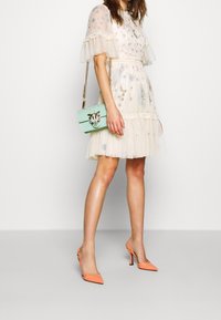 Beige tulleklänning med blomsterbroderi och volangärmar, kombinerad med en mintgrön handväska och orange klackskor.