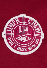 Rød stoff med en sirkulær hvit brodert lapp som viser "UNFR CREW," "UA," og frasen "DON'T MESS WITH US." Teksten inni er fet og fremtredende.