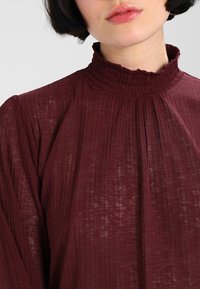 Burgundy paita, jossa on korkea rypytetty kaulus, teksturoitu kangas ja pitkät hihat. Sisältää pystysuoria laskoksia lisäyksinä.