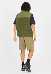 Groene fleece vest met een gladde bovenkant van zwart stof, gecombineerd met khaki shorts. Zwarte wandelschoenen maken de outfit compleet.