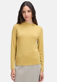 Długo rękawowy top w kolorze musztardowym z wysokim dekoltem i skręconym detalem na ramieniu, wykonany z miękkiego materiału, noszony na wzorzystą spódnicę.
