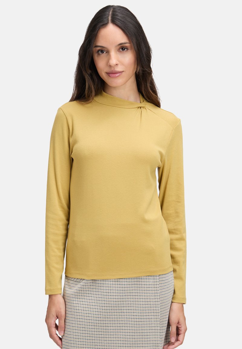 Długo rękawowy top w kolorze musztardowym z wysokim dekoltem i skręconym detalem na ramieniu, wykonany z miękkiego materiału, noszony na wzorzystą spódnicę.