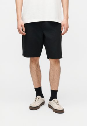Person trägt schwarze knielange Shorts, weiße Turnschuhe mit braunen Sohlen, schwarze Socken und ein weißes T-Shirt und steht vor einem einfarbigen Hintergrund.