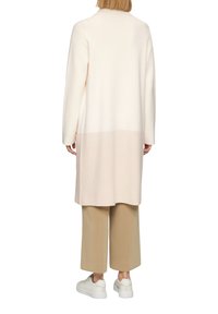Langer, offen gestalteter Cardigan in Hellrosa und Creme. Weiche Textur, gerader Schnitt, mit langen Ärmeln. Kombiniert mit beigen Hosen und weißen Turnschuhen.
