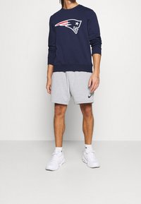 Sweatshirt marine avec le logo des New England Patriots, débardeur, et poignets côtelés, associé à un short gris de Nike. Des chaussures de sport blanches complètent le look.